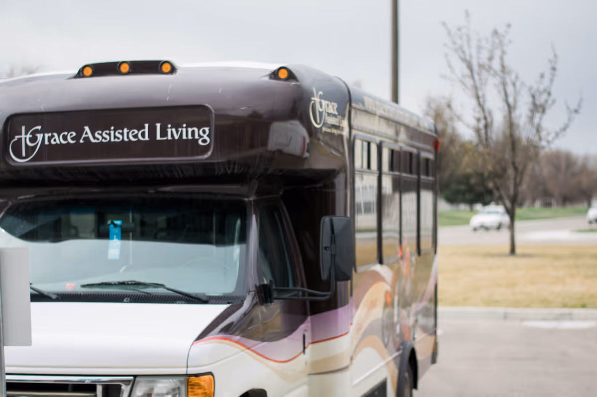 A shuttle bus parked outdoors with the sign 'Grace Assisted Living' displayed on the front above the windshield. The bus is white and brown with a purple and beige wave design on the side. Trees and a grassy area are visible in the background.