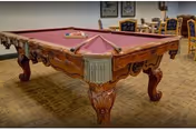 Ornately carved wooden pool table with red felt in a carpeted recreation room with chairs and framed artwork on the walls.