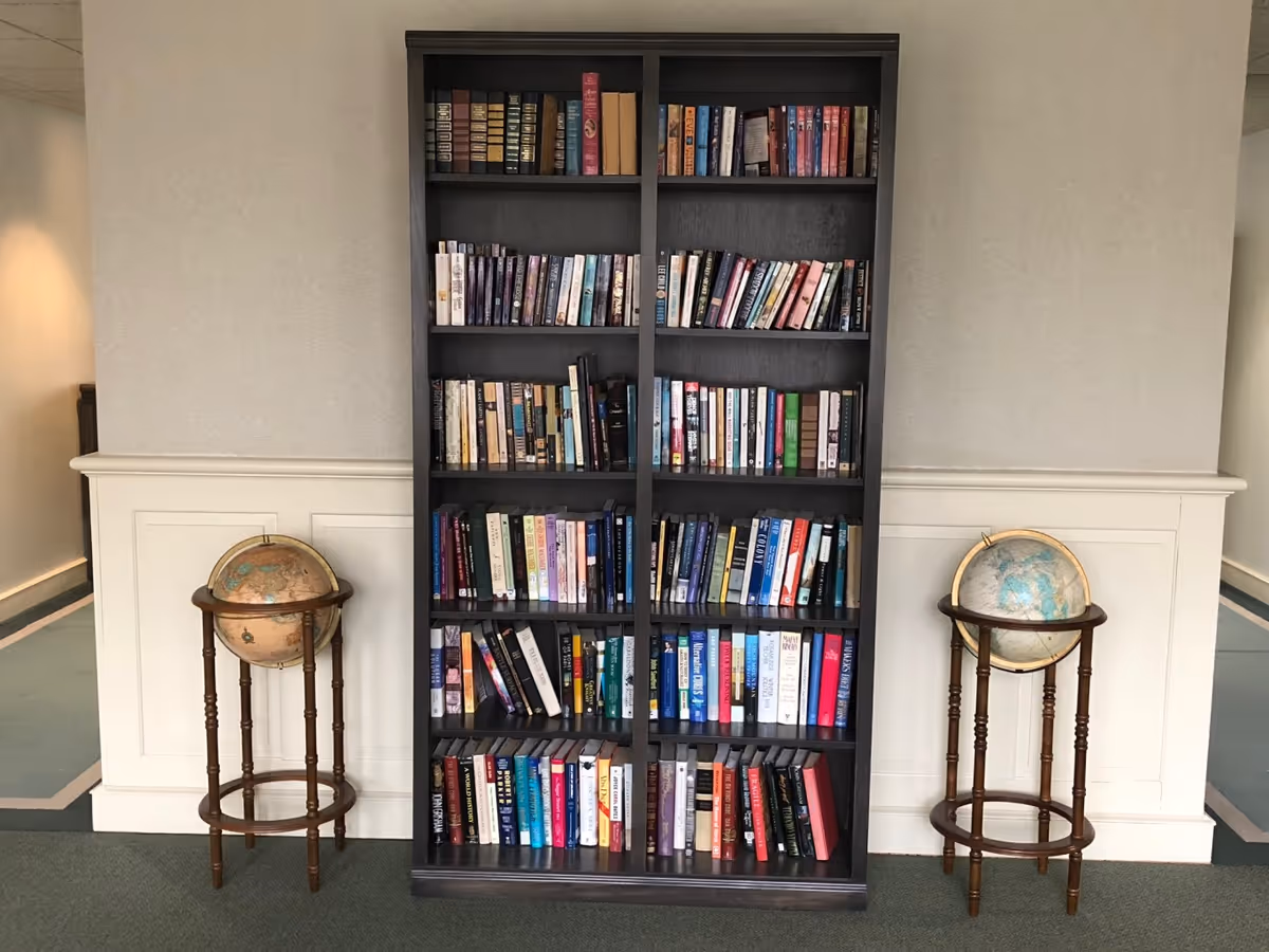 A tall dark wooden bookshelf filled with various books, flanked on each side by a vintage-style globe on a wooden stand, set against a light-colored wall with white wainscoting and green carpeted floor.
