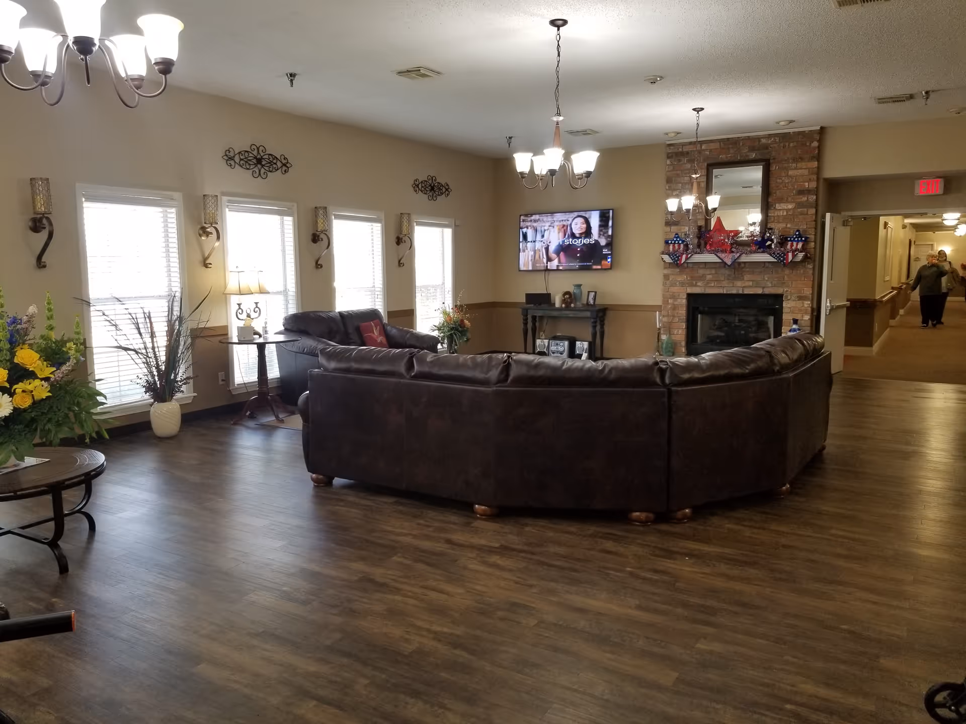 A spacious living room area in Marigold House featuring a large brown leather sectional sofa arranged around a brick fireplace decorated with patriotic star ornaments. The room has multiple windows with blinds, wall sconces, a flat-screen TV mounted on the wall, and several ceiling light fixtures. There are flower arrangements on tables, and a hallway is visible in the background with two people walking.