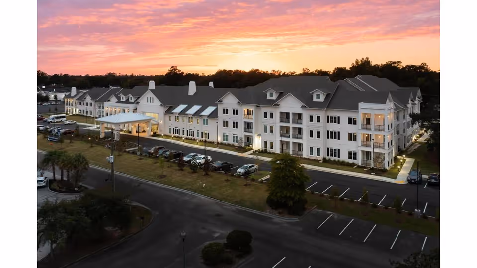 Aerial view of a large white multi-story senior living building with a lit entrance, parking lot, and a colorful sunset sky.