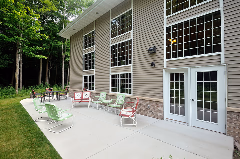 Outdoor patio area at a retirement community with metal chairs and tables on a concrete surface next to a building with large windows and double glass doors, surrounded by green grass and trees.