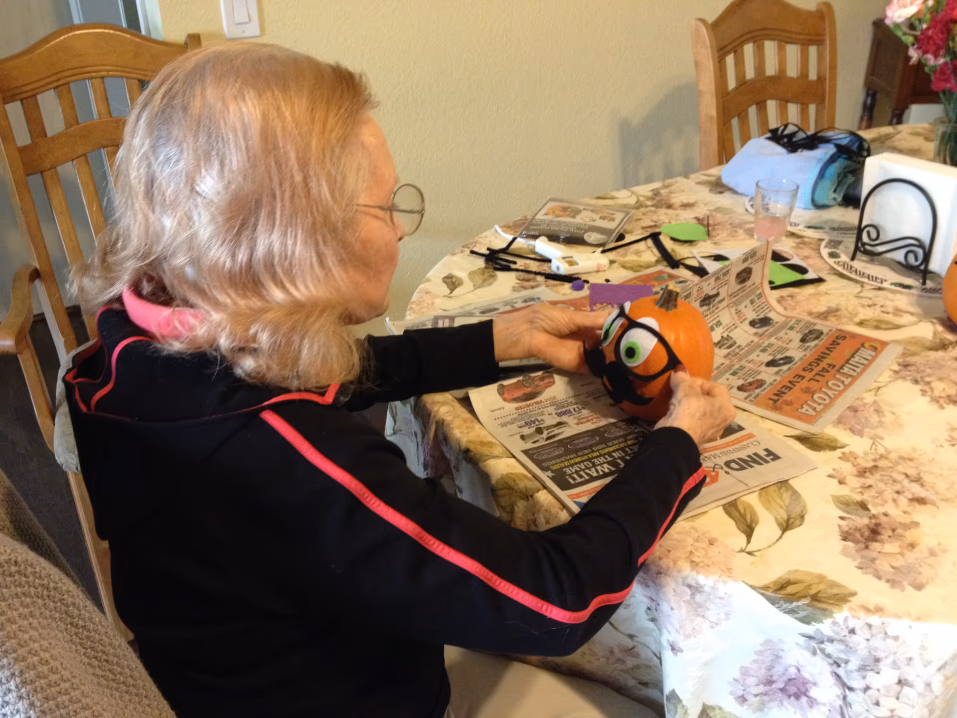 An elderly woman with light hair and glasses is sitting at a dining table covered with a floral tablecloth. She is holding and decorating a small pumpkin with googly eyes and black paper accessories. The table has newspapers, craft supplies, a glass with a pink drink, and a napkin holder.