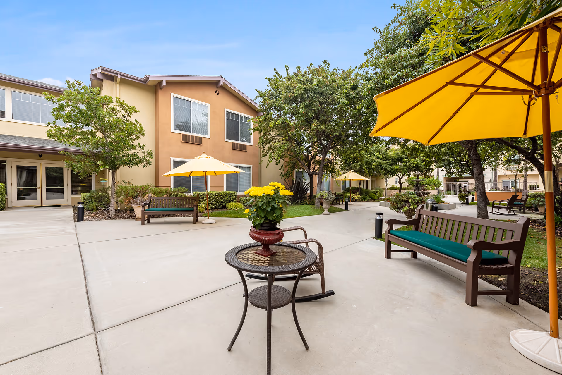 Outdoor courtyard with benches, tables, yellow umbrellas, potted flowers, and a two-story building in the background.