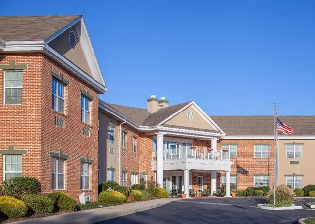 Brick senior living facility exterior featuring a portico entrance, flag, and manicured landscaping.