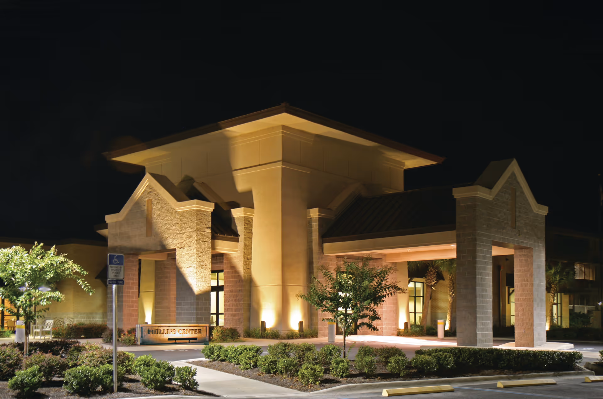 Lit nighttime view of a senior living facility entrance and porte-cochère with landscaping and parking.