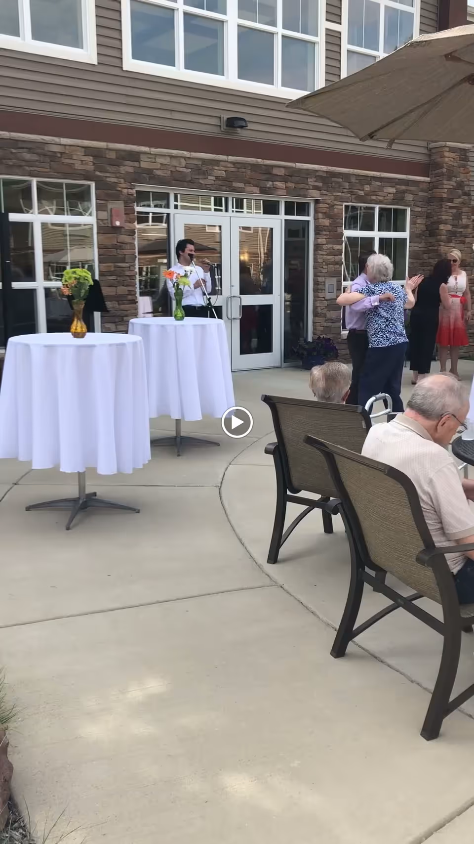 Outdoor patio area at Terra Vista of Oakbrook Terrace with elderly people sitting on chairs and socializing. Two tall round tables with white tablecloths and flower vases are visible. A man is playing a musical instrument near the building entrance, and two women are hugging. The building has stone and siding exterior with large windows and a door.