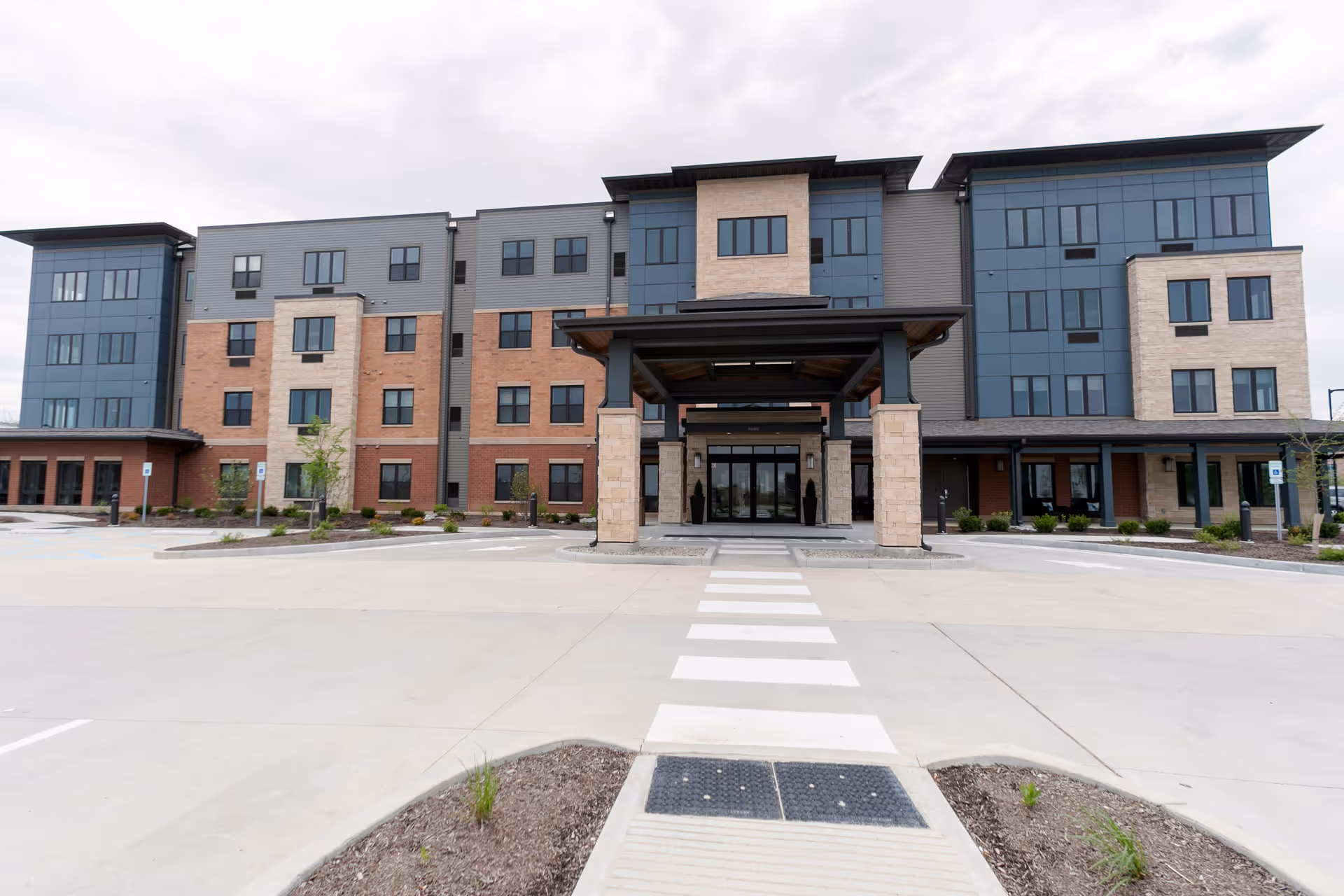Front exterior view of a modern multi-story senior living facility building with a covered entrance, large windows, and a paved driveway with a pedestrian crosswalk leading to the entrance.