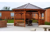 A wooden gazebo with a metal roof situated on a concrete patio area in front of a brick building. There are tables and chairs inside the gazebo and a stone birdbath or table in the foreground. The scene is outdoors with grass and some trees in the background.