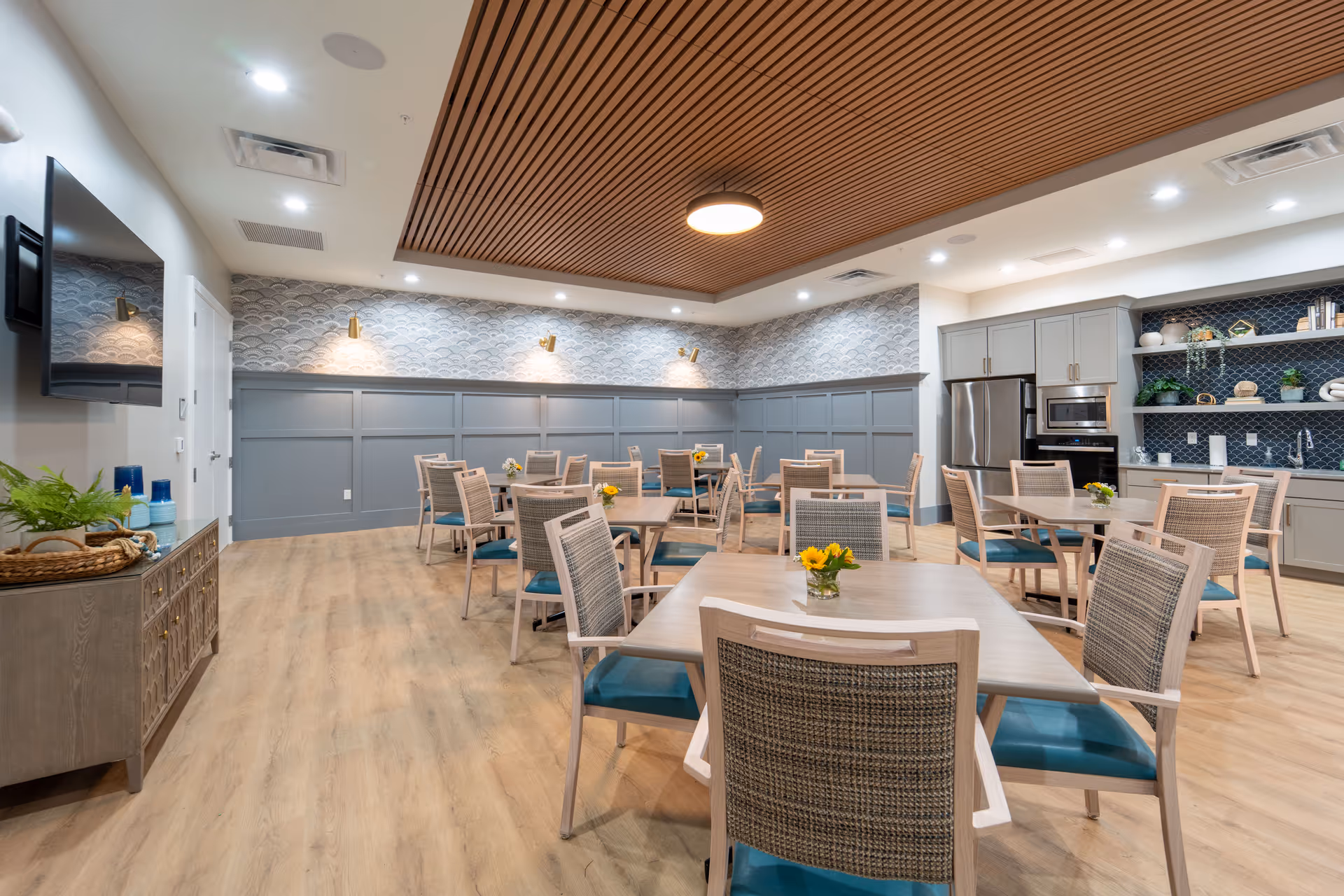 A modern dining room with several wooden tables and chairs with teal cushions. The room features a wooden slatted ceiling panel with a round light fixture, light wood flooring, and a gray wainscoted wall with patterned wallpaper above. On the right side, there is a kitchenette area with gray cabinets, a stainless steel refrigerator, microwave, and open shelves with decorative items. A flat-screen TV is mounted on the left wall above a wooden sideboard with plants and decorative items.