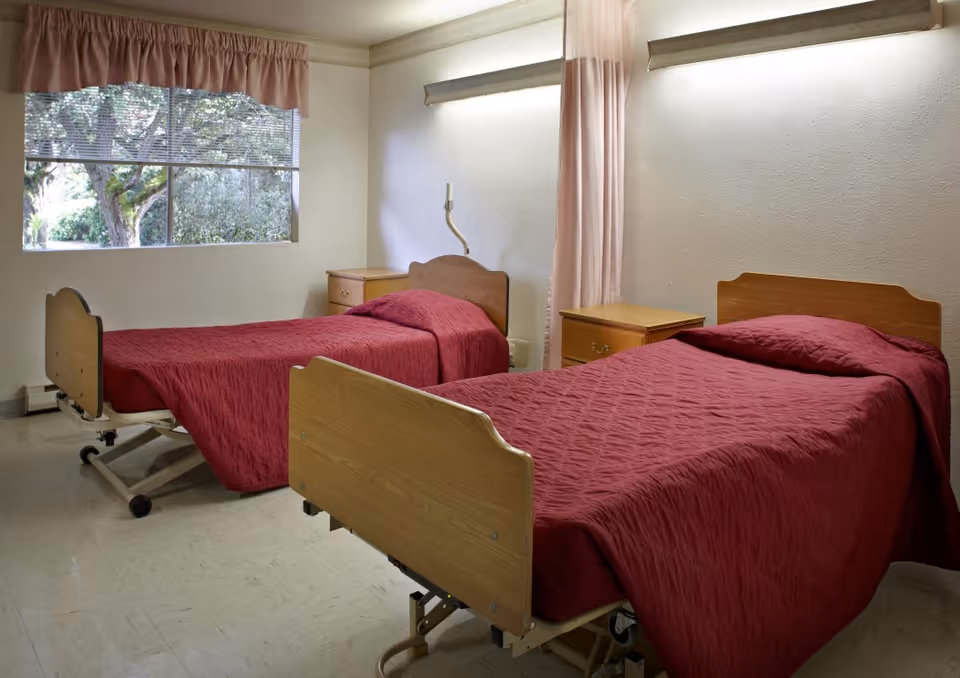 A room with two hospital-style beds covered in red bedspreads, each with a wooden headboard and footboard. There are two wooden nightstands between and beside the beds. A large window with blinds and a pink valance lets in natural light, and a pink privacy curtain hangs between the beds.