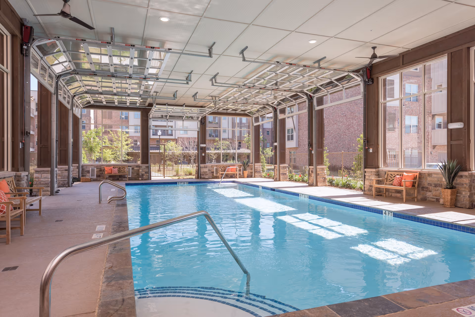 Indoor swimming pool area with clear blue water, surrounded by large windows and stone walls. There are wooden benches with orange cushions along the sides, ceiling fans, and a partially open garage-style door letting in natural light. Outside, a garden and brick buildings are visible.