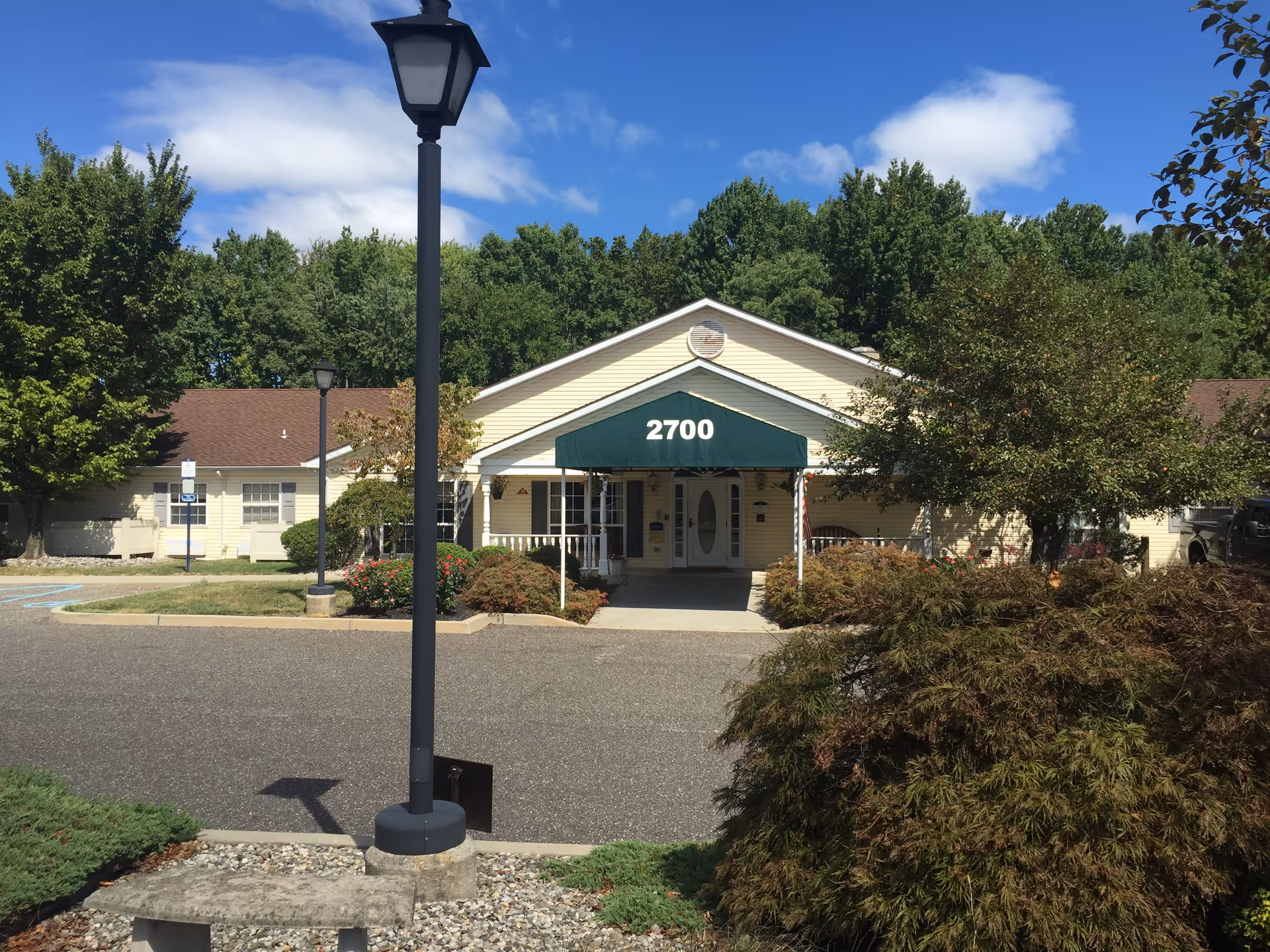 Front exterior view of a single-story building with a green awning displaying the number 2700. The building is surrounded by trees, bushes, and a parking area with a lamp post in the foreground under a partly cloudy sky.