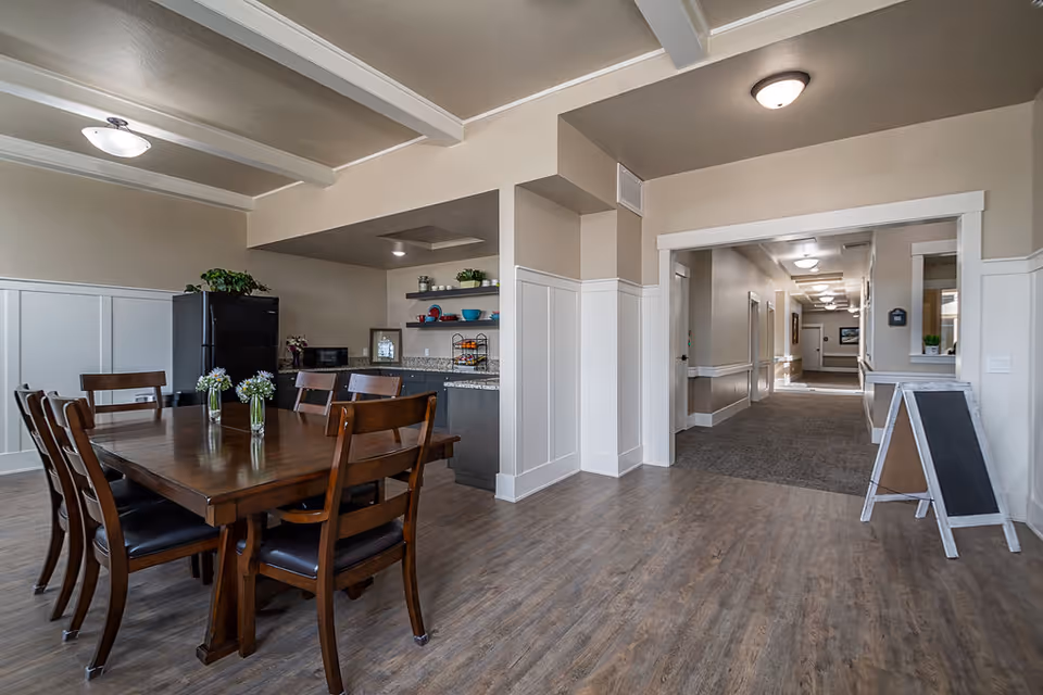 A dining area in a senior living facility with a wooden table and six chairs. The table has three small vases with flowers. In the background, there is a kitchenette with a black refrigerator, microwave, and open shelves holding dishes and plants. To the right, a hallway leads to other rooms, and a small chalkboard sign is placed near the entrance to the hallway. The room has wood flooring, beige walls with white wainscoting, and ceiling beams.
