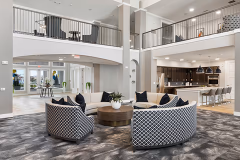 Spacious and modern senior living facility common area with a circular seating arrangement featuring patterned armchairs and white sofas around a round wooden coffee table. The room has a high ceiling with a second-floor balcony overlooking the area. In the background, there is a kitchen with a large island and bar stools, and large windows letting in natural light.