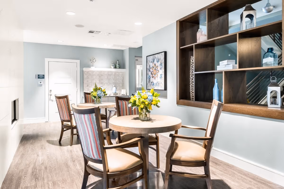 A bright and modern dining area with round wooden tables and chairs featuring striped upholstery. Each table has a vase with yellow and blue flowers. The room has light blue walls, a wooden shelving unit with decorative items, a framed floral artwork, and a door in the background.