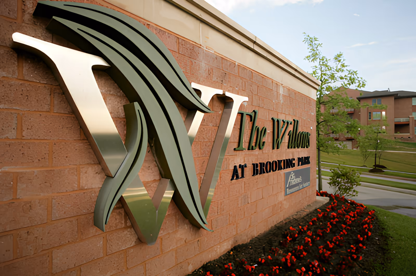 Decorative metal sign on a brick wall reading 'The Willows at Brooking Park' with red flowers and the facility buildings and lawn visible in the background.