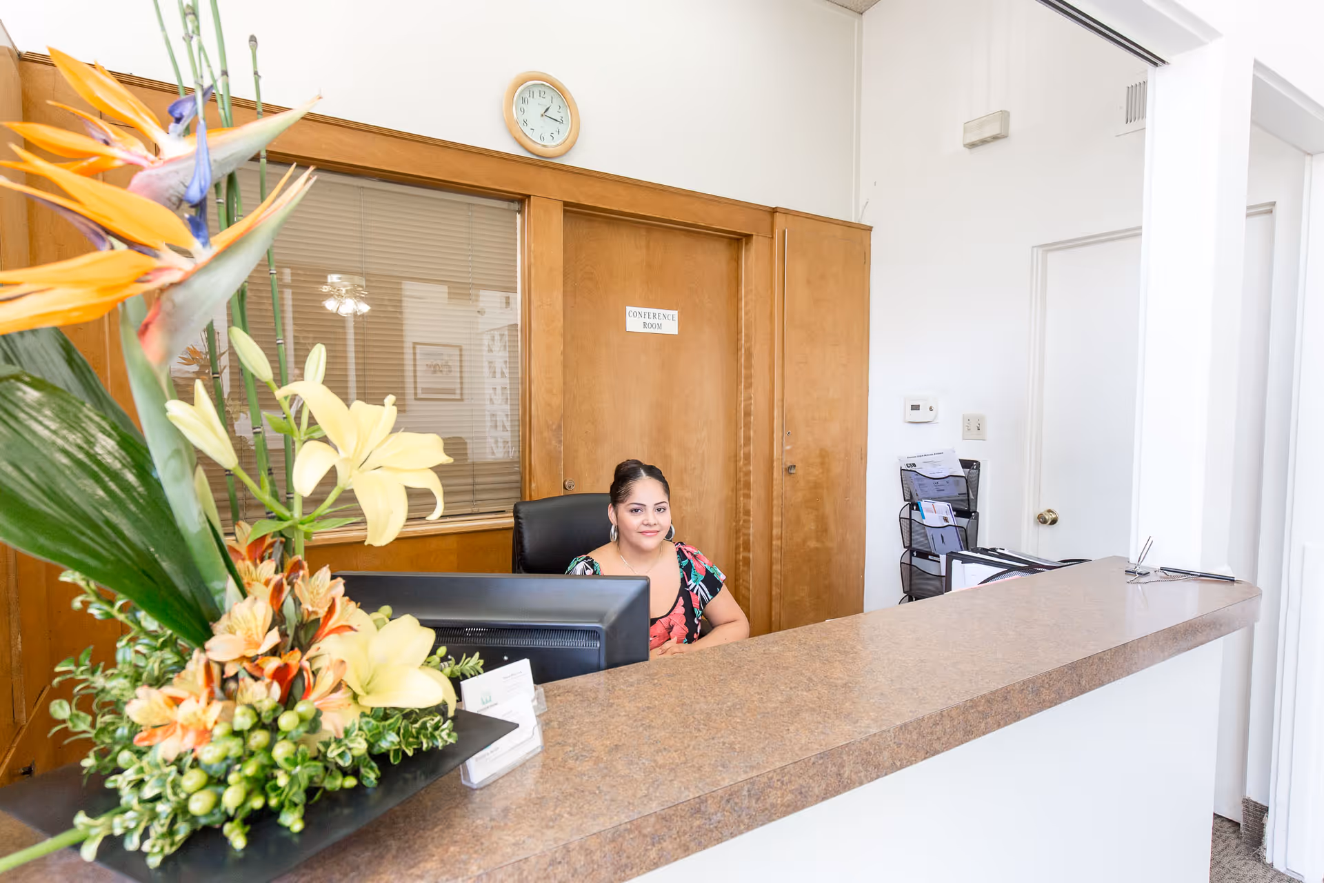 Reception area with a woman sitting behind a desk with a computer monitor. There is a large floral arrangement on the counter. Behind her is a wooden door labeled 'Conference Room' and a window with blinds. A clock is mounted above the door on the wall.