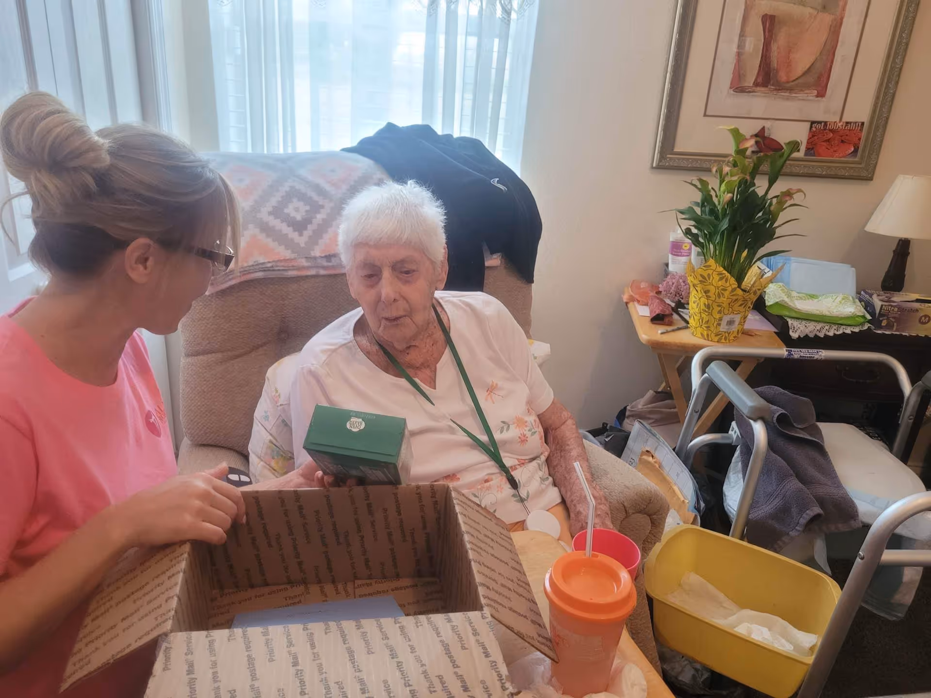 An elderly woman sitting in a cushioned armchair holding a small green box, with a younger woman in a pink shirt sitting beside her holding an open cardboard box. The room has a side table with a potted plant, various items, a walker, and a yellow waste bin. There is a window with sheer curtains behind them.