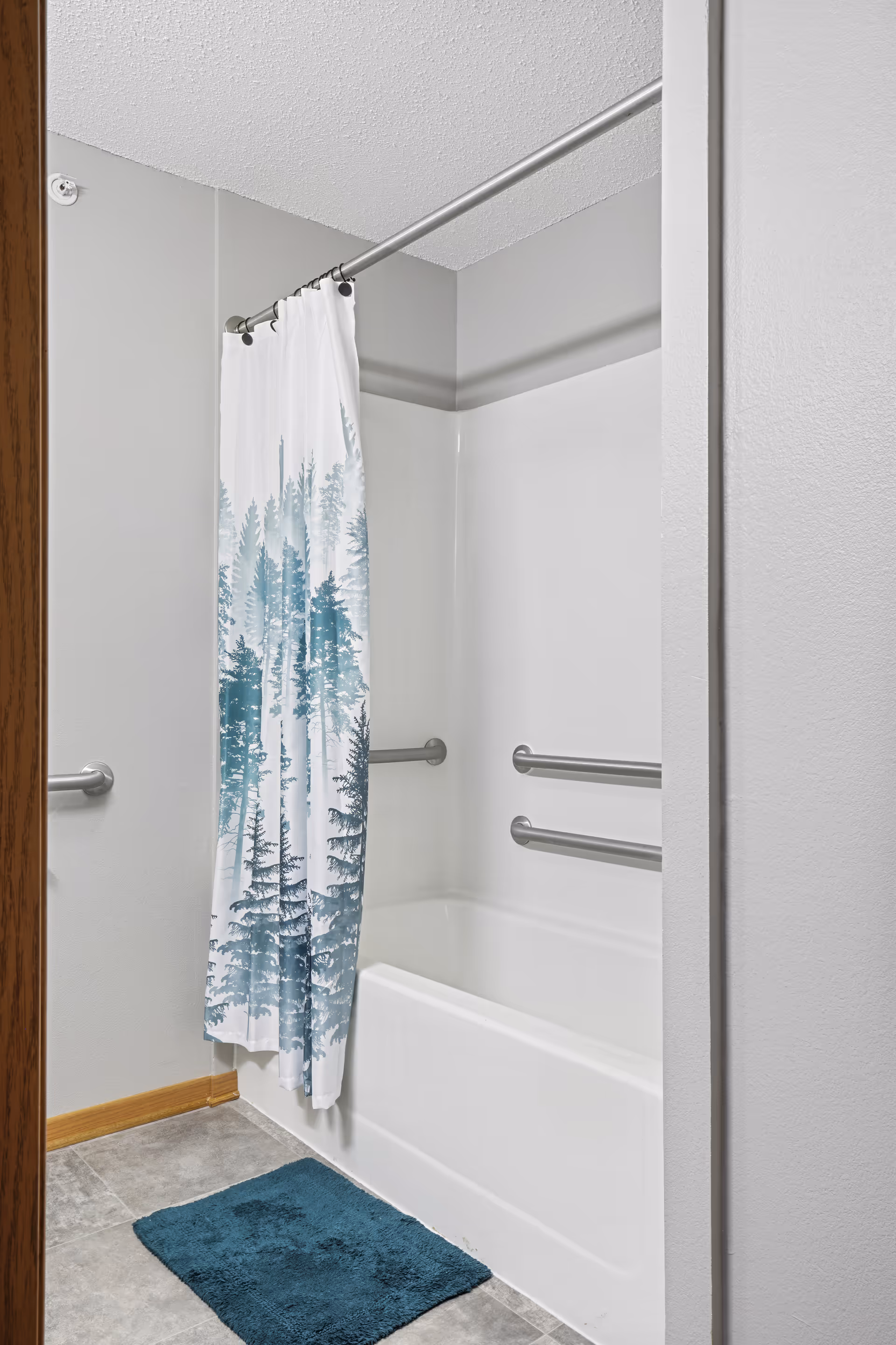 Bathtub and shower with a blue tree-patterned curtain, wall-mounted grab bars, and a teal bath mat on a tiled floor.