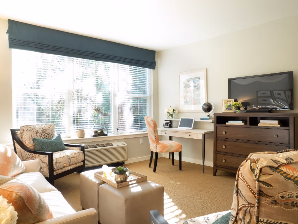 Sunlit living room with a large window, armchairs, ottomans, a desk and chair, and a TV on a dresser.