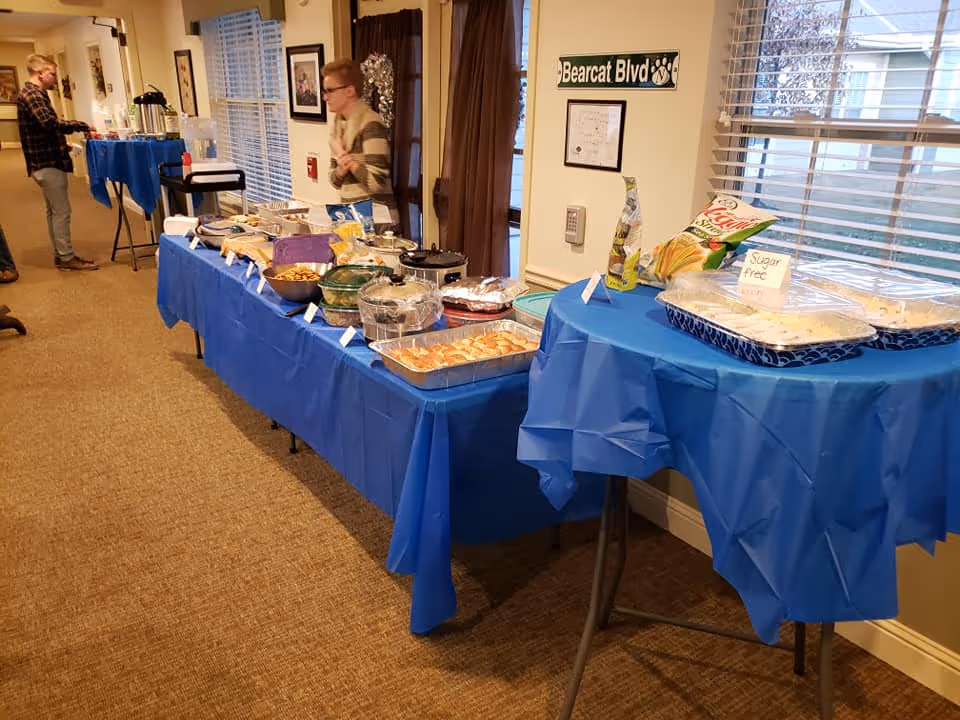 A buffet setup with various dishes covered and labeled on tables draped with blue tablecloths in a room with carpeted floor and windows. Two people are standing near the buffet, one serving themselves food. A sign on the wall reads 'Bearcat Blvd'.