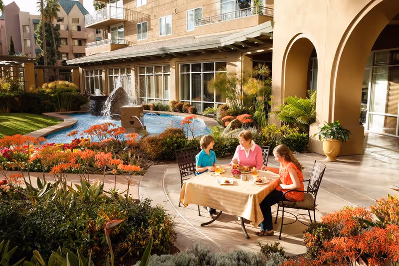 Three people sitting at a table with a beige tablecloth outdoors near a water fountain surrounded by colorful flowers and greenery, with a building featuring large windows and arches in the background.