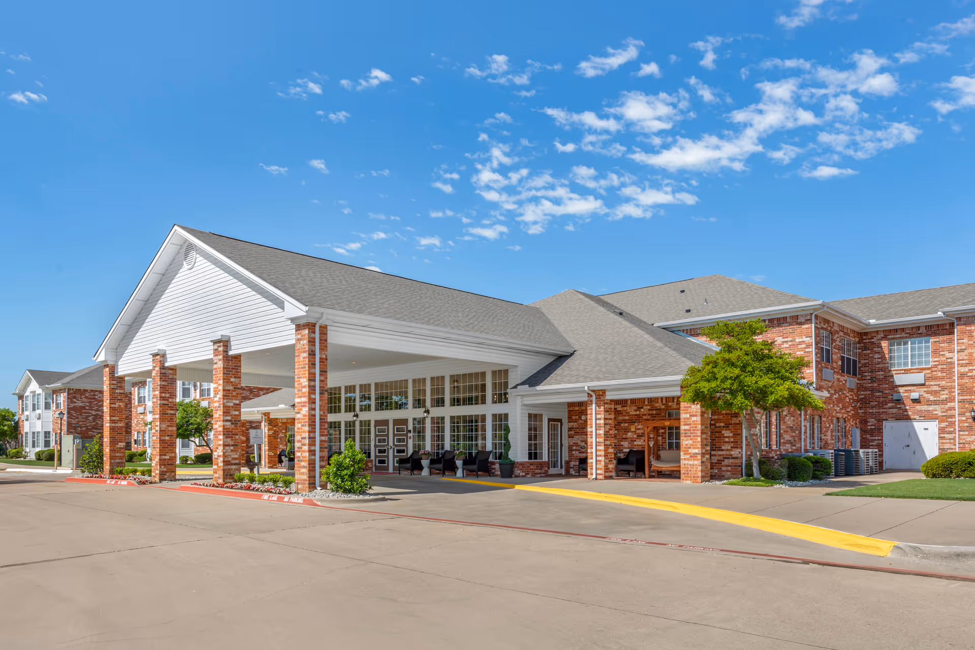 Front entrance of a brick senior living facility with a covered porte-cochère and seating under a clear blue sky.