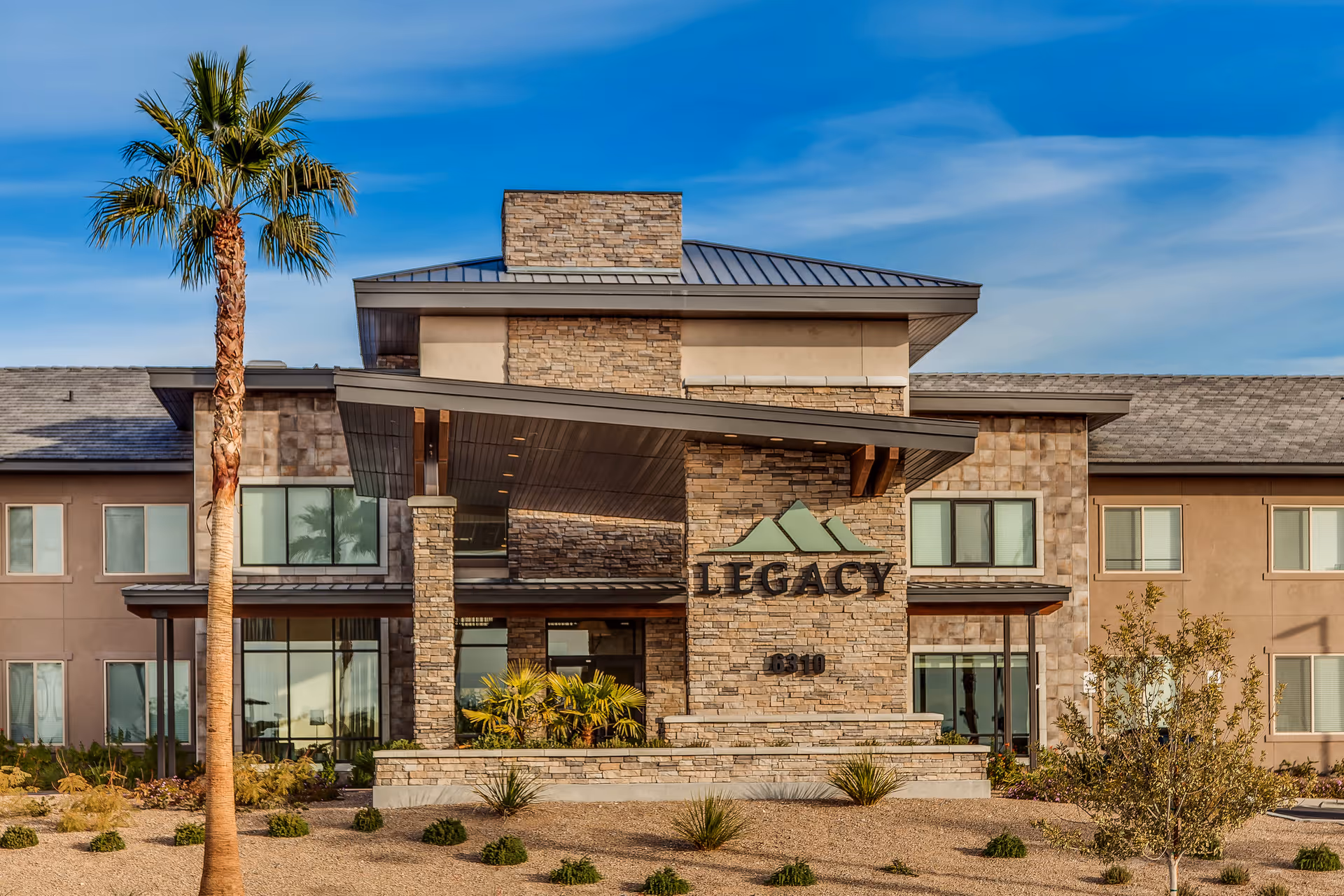 Front exterior view of Legacy House of Centennial Hills building with stone and beige facade, large windows, a covered entrance, and desert landscaping including a palm tree and small shrubs under a blue sky.