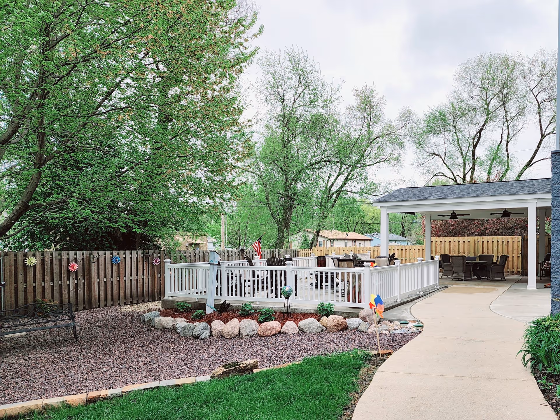 Outdoor patio area at Springfield Supportive Living Center featuring a white fenced deck with seating, a covered seating area with tables and chairs, a curved concrete walkway, green grass, trees, and a wooden fence decorated with colorful flower ornaments.
