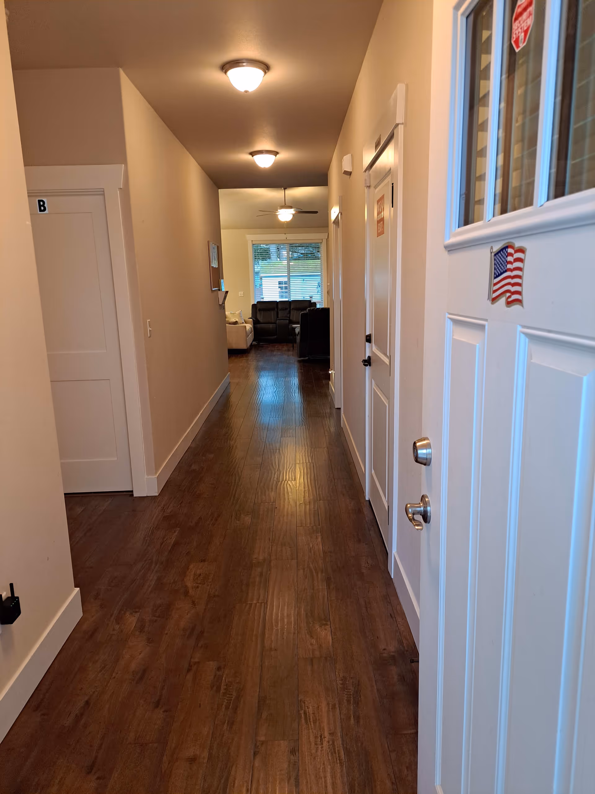 A long hallway with wooden flooring and beige walls leading to a living room area with sofas and a window. The hallway has ceiling lights and doors on both sides, one door marked with a letter B and another with a sign. The front door is partially open and has an American flag sticker on it.