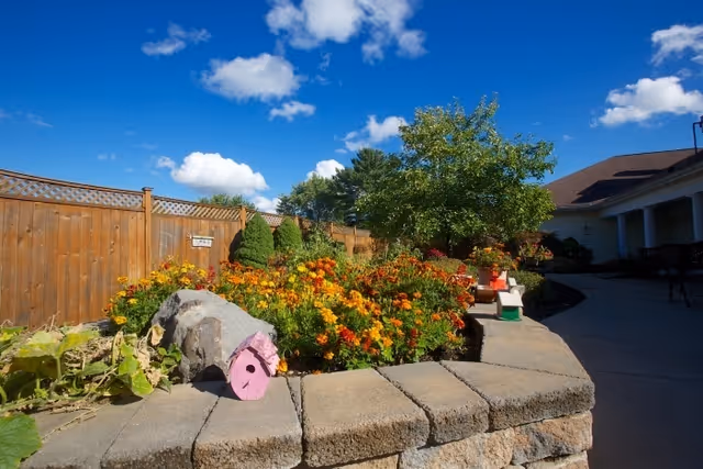 Sunny outdoor courtyard with a stone planter full of colorful flowers, a wooden fence, and a paved walkway alongside a building.