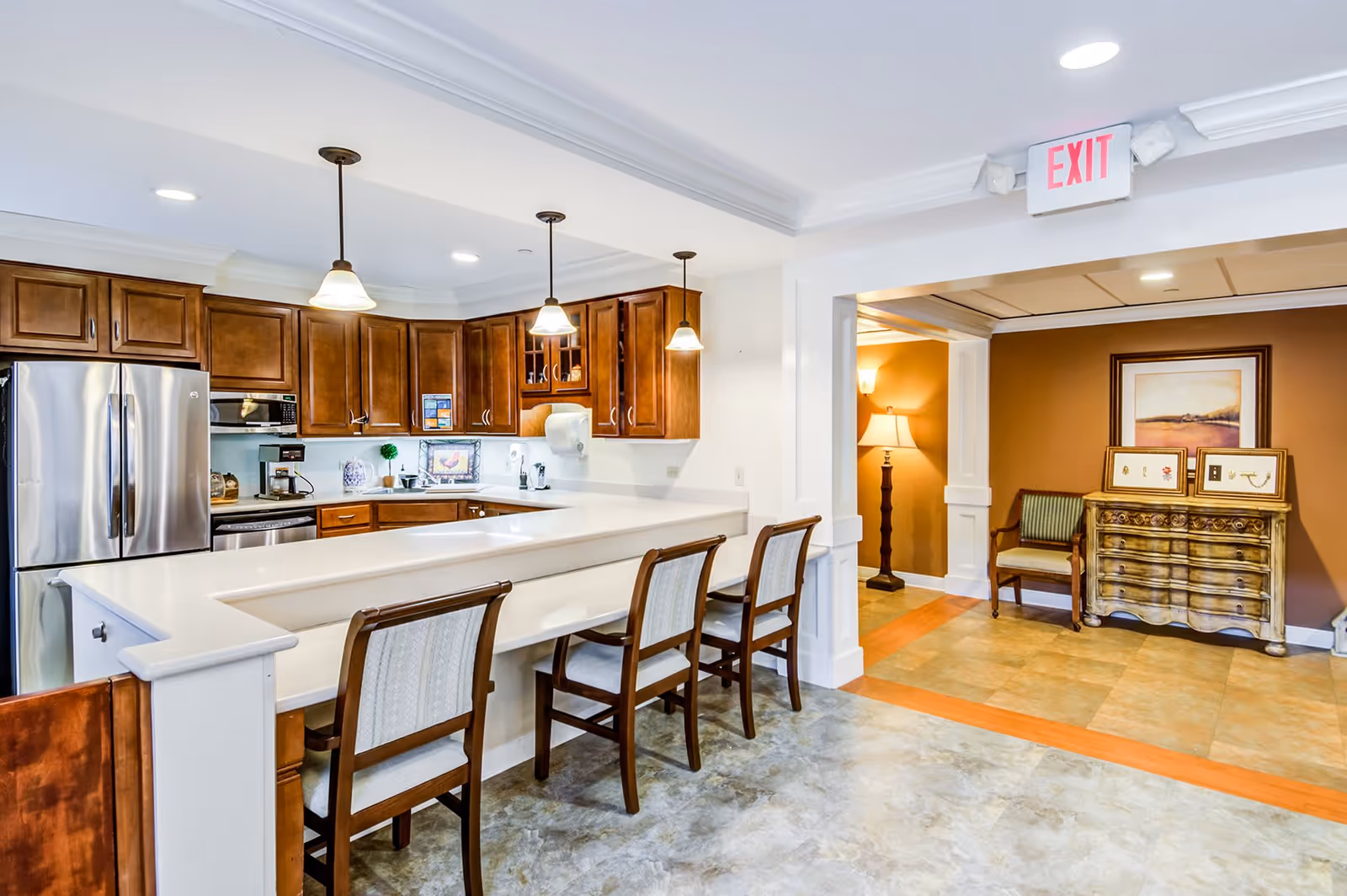 A bright, modern kitchen area with wooden cabinets, stainless steel refrigerator, and a white countertop bar with four wooden chairs. The kitchen opens into a hallway with a decorative wooden chest, framed artwork, a green chair, and a floor lamp. An exit sign is visible on the ceiling.