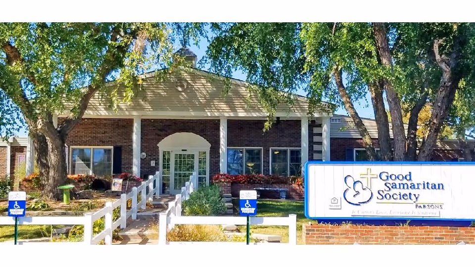 Front entrance of the Good Samaritan Society - Parsons building with a white fence, large sign on the lawn, and trees framing the doorway.