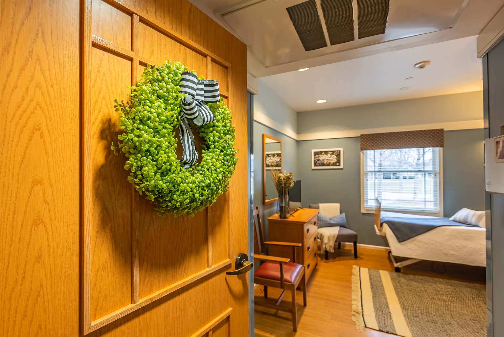 View into a cozy bedroom in a senior living facility with a wooden door decorated with a green wreath and striped bow. Inside the room, there is a bed with white and blue bedding near a window with blinds and a valance. A wooden dresser with a vase of flowers and a mirror above it is against the wall, along with a chair and a small table. The room has wooden flooring and a rug.