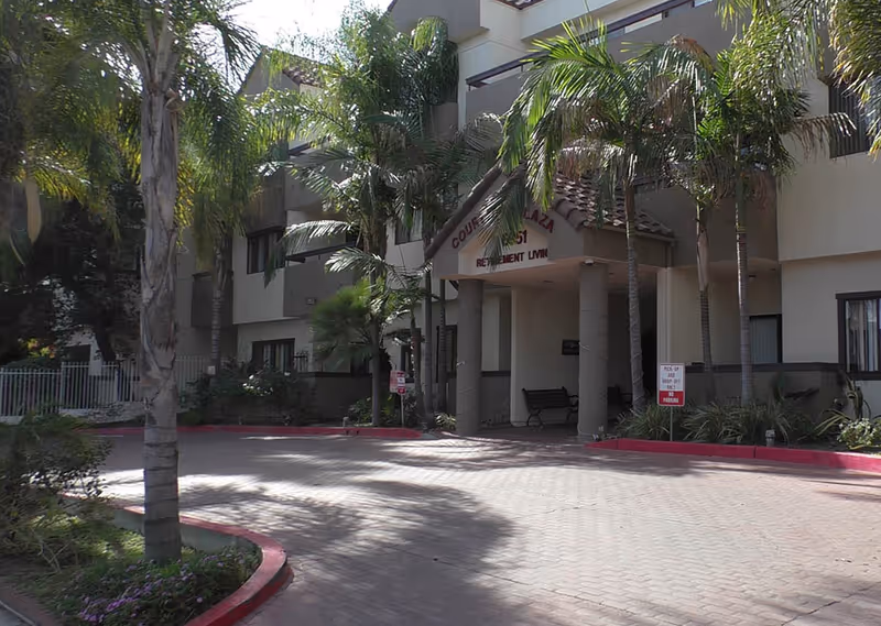 Entrance of a retirement living facility named Court Plaza with palm trees and a paved driveway leading to the covered entrance supported by columns.