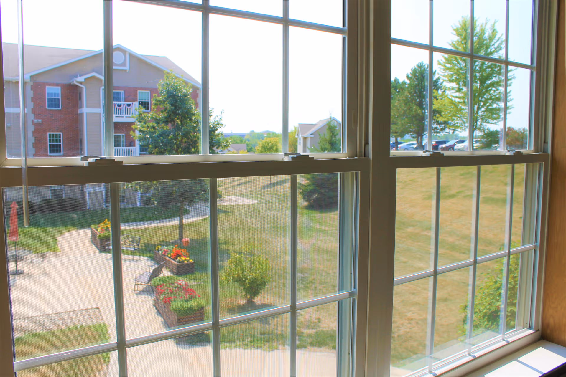View through a multi-pane window of a landscaped courtyard with flower planters, benches, walking paths, and a brick building.