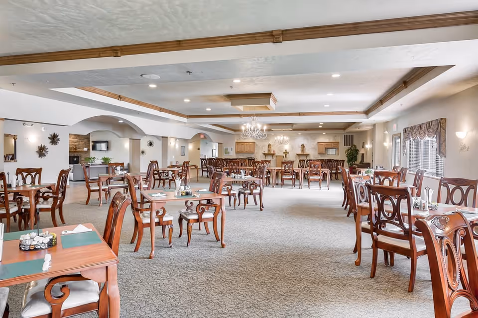 Spacious senior dining room with multiple wooden tables and chairs, chandeliers, and windows along the right wall.