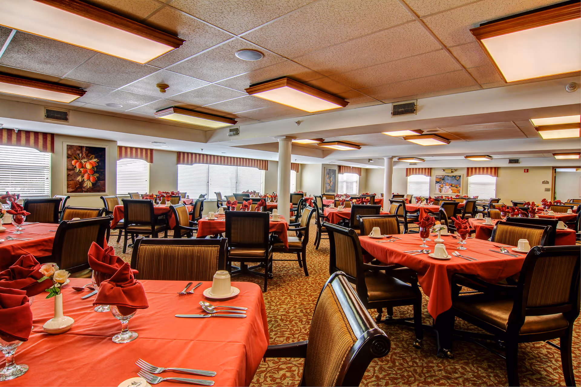 A spacious dining room with multiple tables covered in red tablecloths, each set with folded red napkins, glassware, silverware, and white cups. The room has patterned carpet flooring, several windows with striped valances, and framed artwork on the walls. The ceiling has recessed lighting fixtures.
