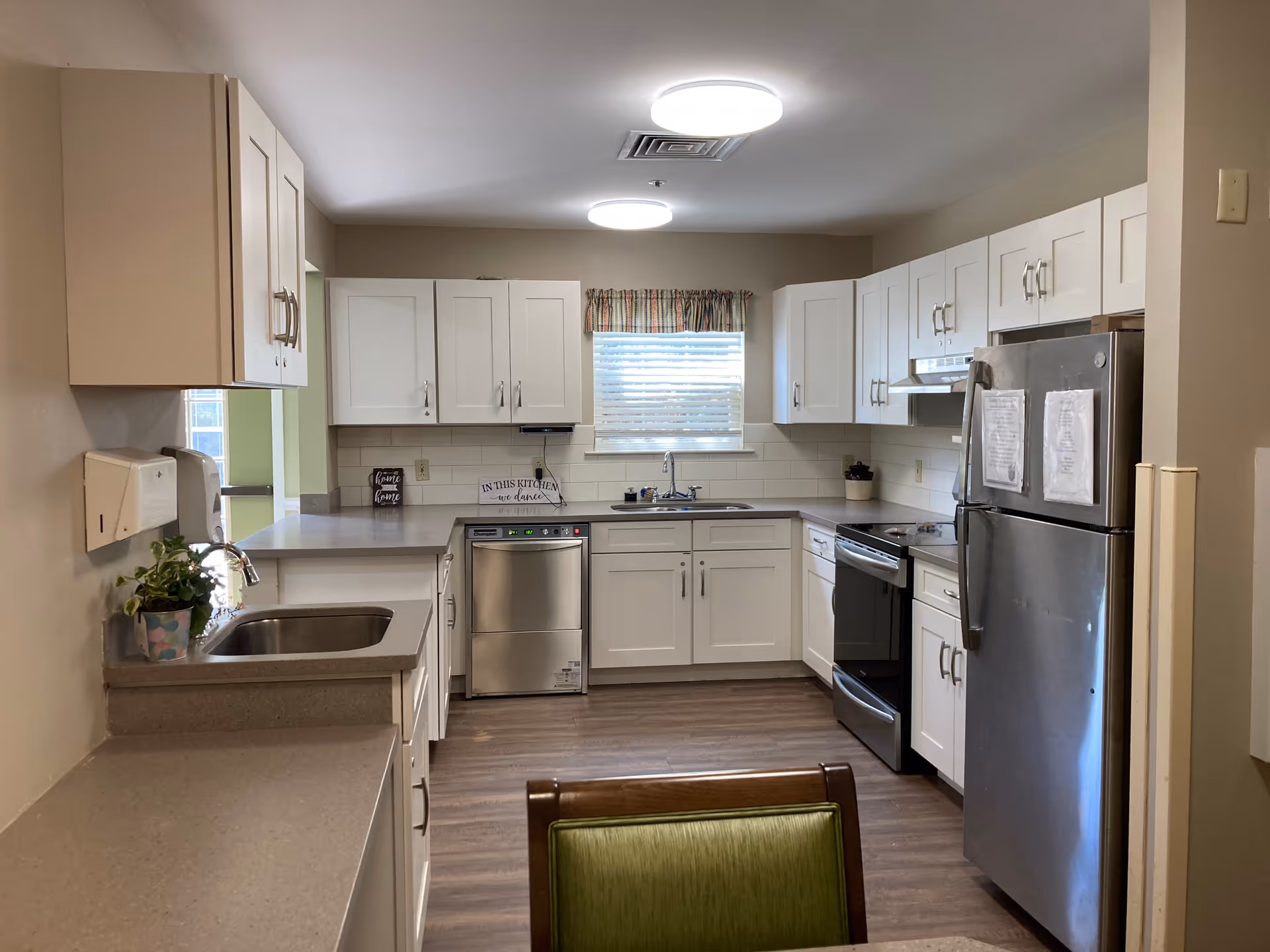 A clean, well-lit kitchen with white cabinets, stainless steel appliances including a dishwasher, stove, and refrigerator. There is a window with blinds and a colorful valance above the sink. The countertops are beige, and the floor is wood. A green cushioned chair is partially visible in the foreground.