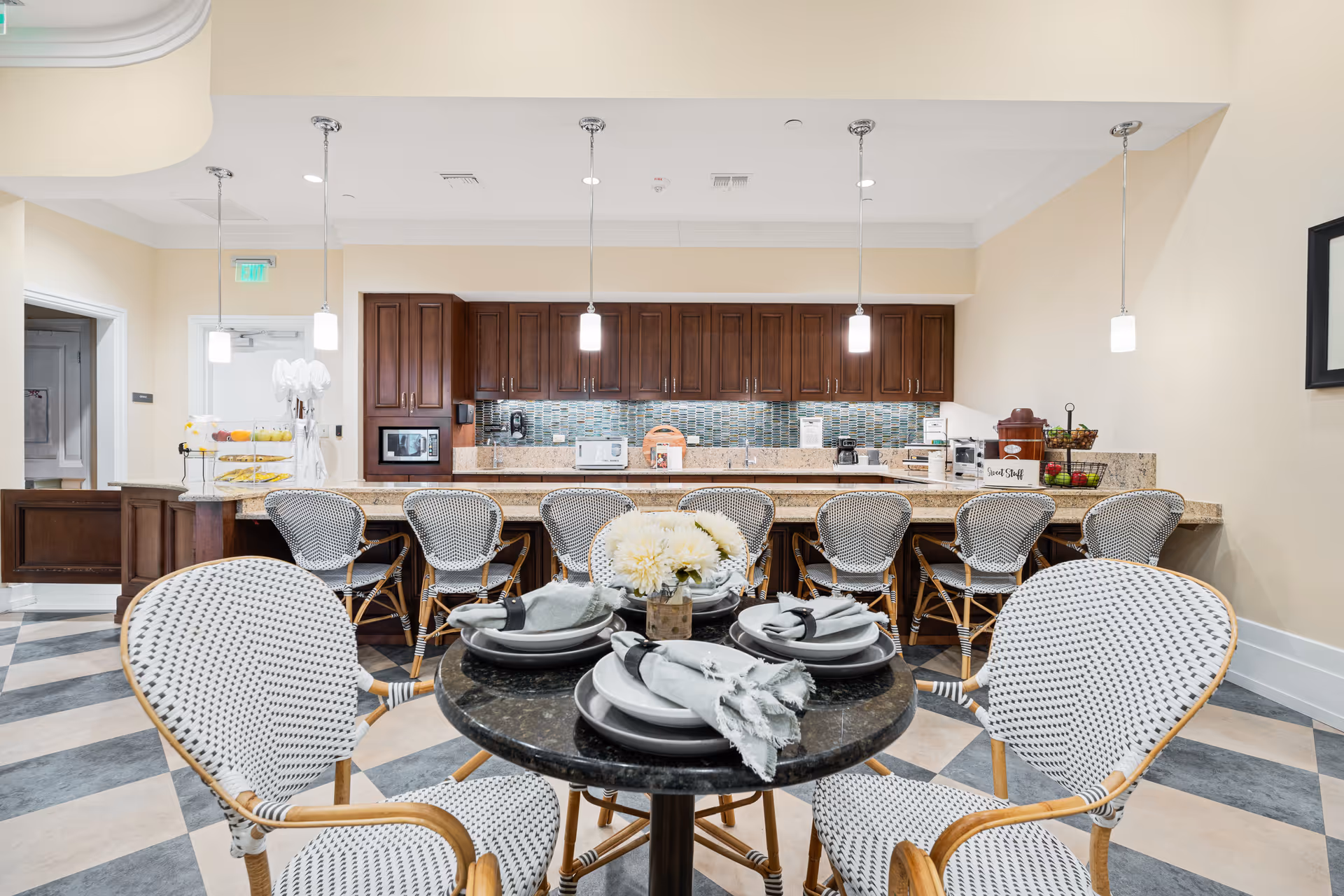 A bright and clean dining area in a senior living facility featuring a round table set with plates, napkins, and a flower centerpiece in the foreground. Behind the table is a long counter with several wicker chairs and a kitchen area with wooden cabinets, a microwave, and various kitchen appliances. The floor has a checkered pattern, and pendant lights hang from the ceiling.