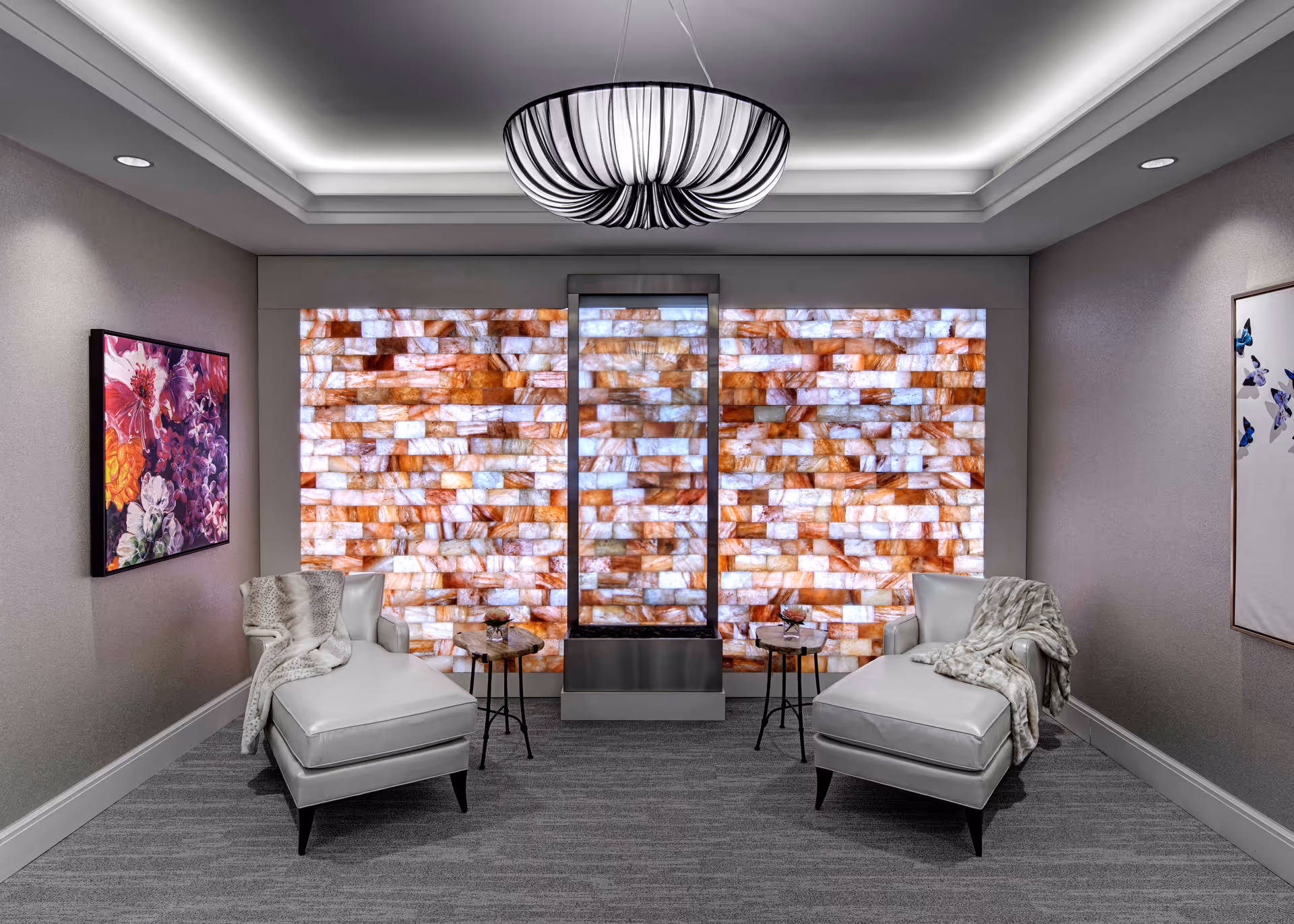 A modern lounge area with two white chaise lounge chairs draped with light gray blankets facing a decorative wall made of illuminated rectangular stone tiles in shades of white, orange, and brown. Between the chairs are two small round side tables with decorative items. The room has gray carpet, neutral-colored walls with floral artwork on each side, and a contemporary black and white pendant light hanging from the ceiling.