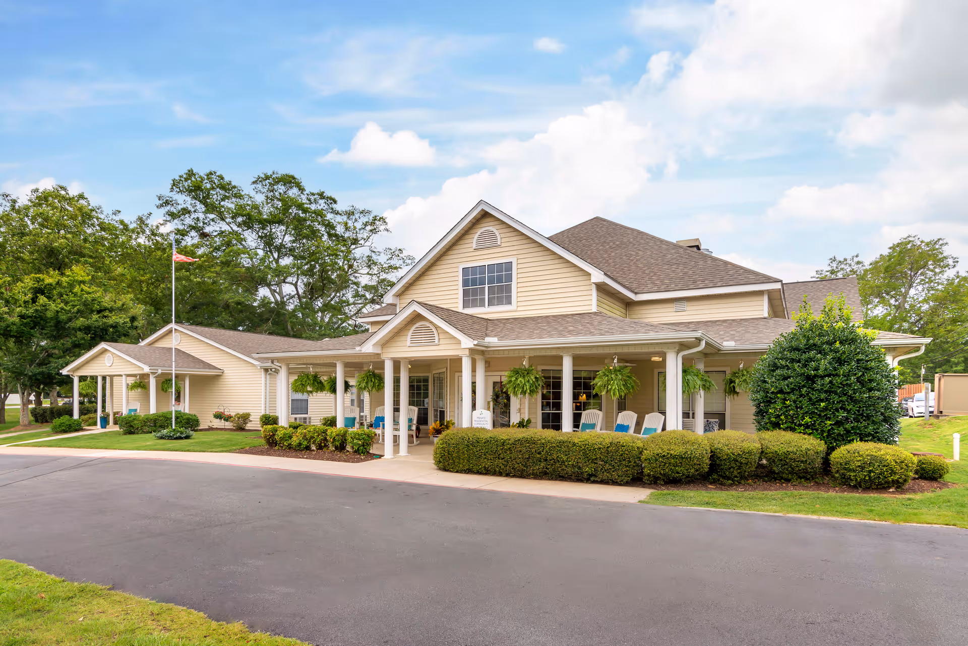 Exterior view of a single-story senior living facility building with beige siding and a brown roof. The building has a covered porch with hanging plants and several chairs. There are bushes and a small lawn in front, with a paved driveway leading up to the entrance. Trees and a partly cloudy sky are visible in the background.