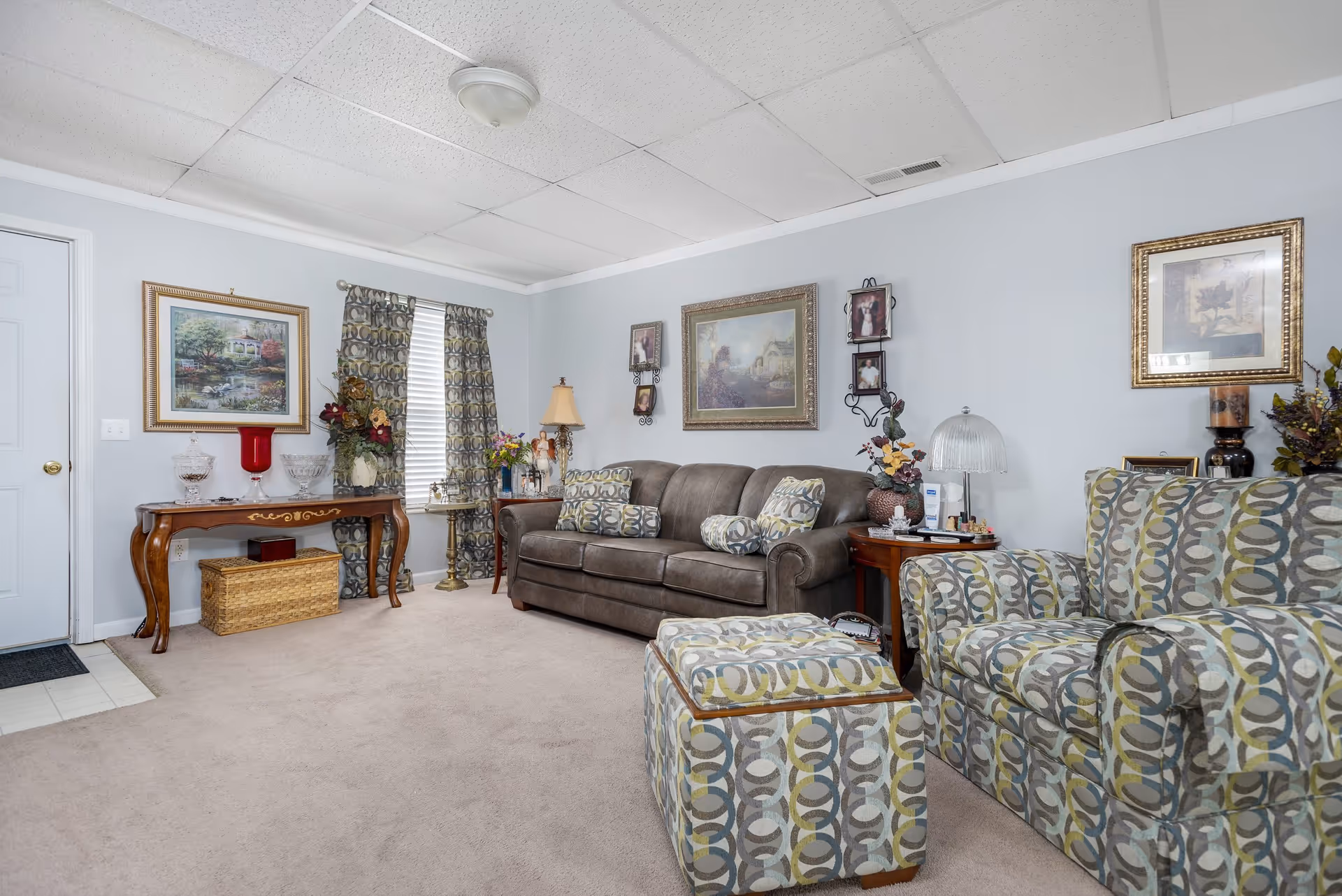 A cozy living room with a brown leather sofa adorned with patterned cushions, a matching patterned armchair and ottoman, and wooden side tables with decorative lamps and flowers. The walls are light blue with framed artwork and photos, and there is a window with patterned curtains. A wooden console table with glassware and a woven basket underneath is positioned against one wall.