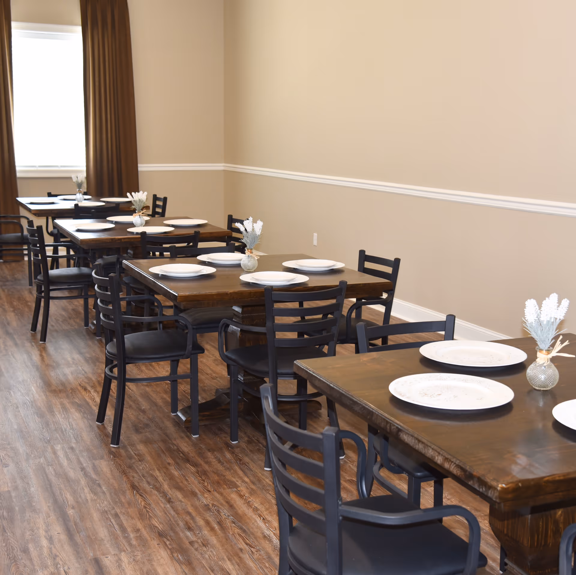 A dining room with several wooden tables set with white plates and small vases containing white flowers. The room has wooden flooring, beige walls with white trim, and dark brown curtains covering a window.