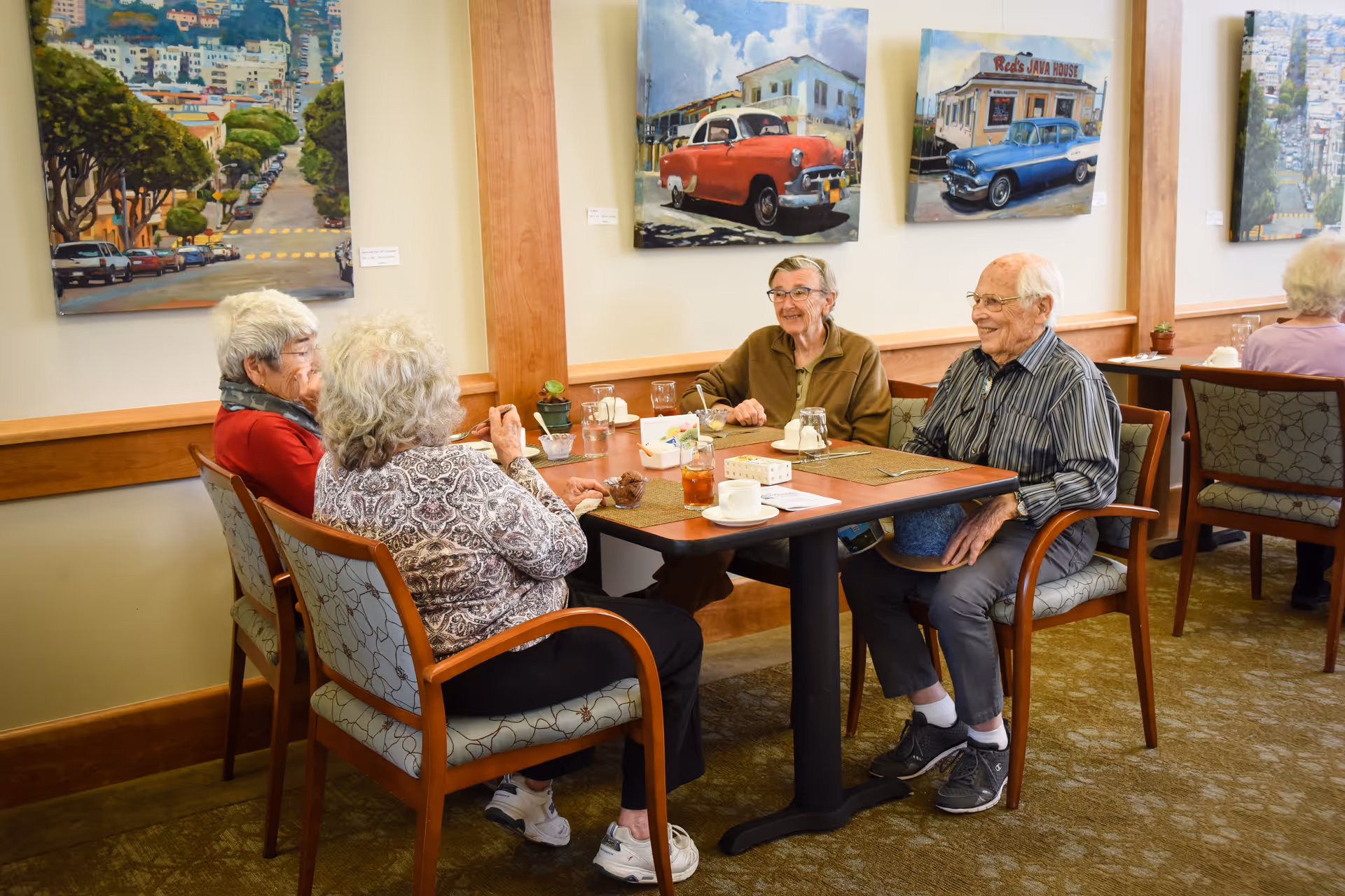 Four elderly people sitting around a dining table in a well-lit room, enjoying conversation and drinks. The walls are decorated with colorful paintings of vintage cars and cityscapes. The room has wooden trim and carpeted floors.
