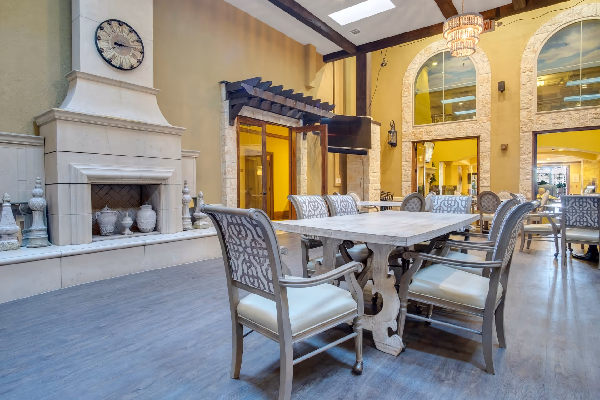 A bright communal dining area with a large wooden table surrounded by upholstered chairs, a decorative fireplace, and high ceilings with skylights and a chandelier.