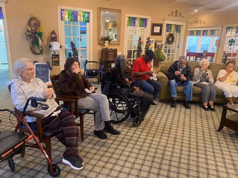 A group of elderly people sitting in a common living area of a senior living facility, some seated on chairs and others on a couch, enjoying snacks. The room is decorated with colorful Mardi Gras-themed decorations, including wreaths and garlands on the windows and walls. There is a walker visible next to one of the seated individuals.