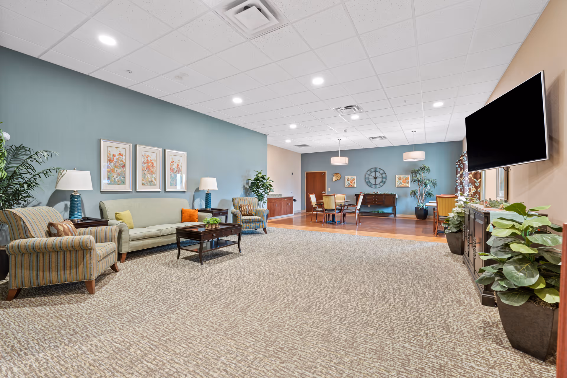 A spacious senior living facility common area with a seating arrangement including a green sofa, two striped armchairs, and a wooden coffee table. The walls are painted in soft green and beige tones, decorated with framed floral artwork and a large clock. There are several potted plants, a flat-screen TV mounted on the wall, and a dining area with a table and chairs in the background.