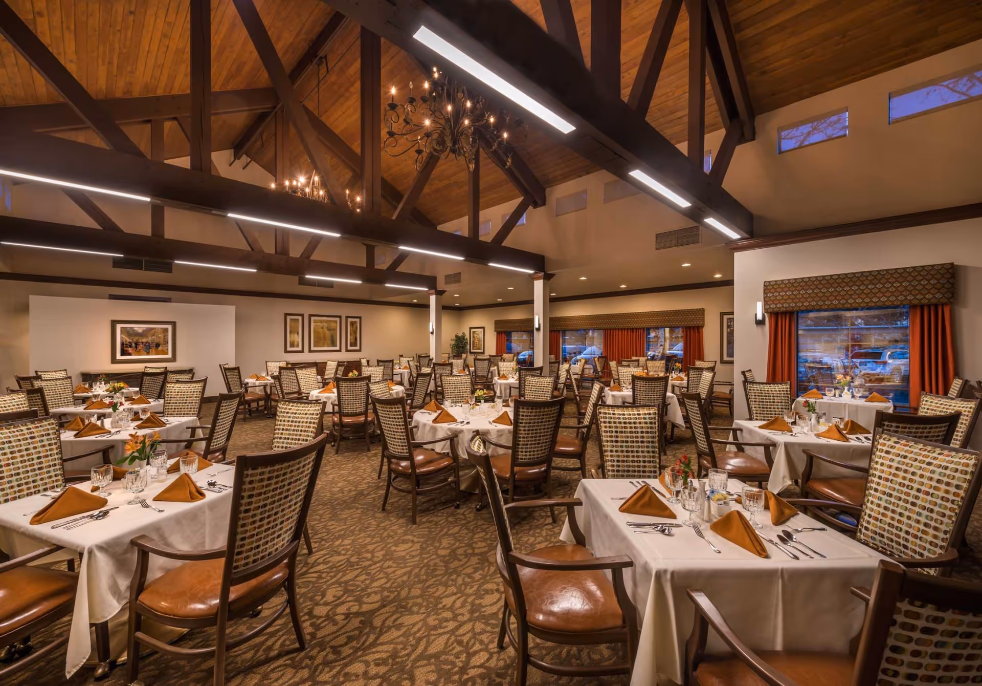 A spacious dining room with multiple tables covered in white tablecloths, each set with brown folded napkins, glassware, and silverware. The room features high wooden ceilings with exposed beams and chandeliers, patterned carpet, and large windows with red curtains.